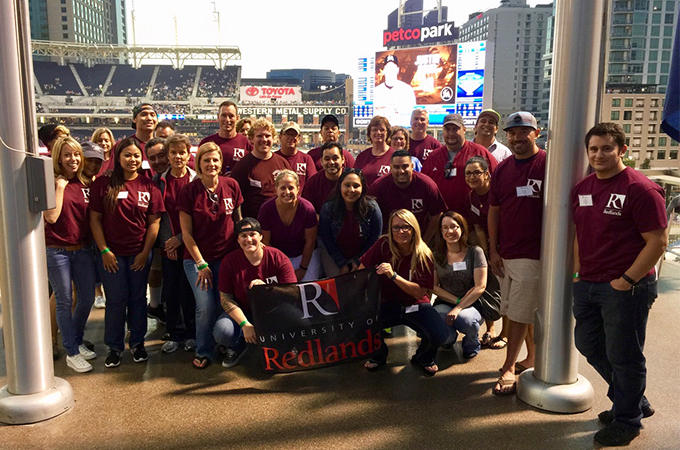 A group of U of R alumni pose together with a U of R flag at a baseball stadium.